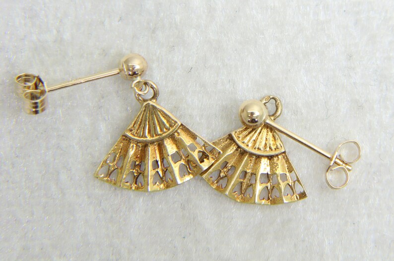 May include: A pair of gold-colored fan-shaped earrings. Each earring features a detailed, openwork design with a post and a small ball at the top. The earrings are displayed on a light-colored surface.