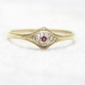 May include: A gold ring featuring a central red gemstone surrounded by six small clear stones arranged in a cluster. The setting is shaped like a diamond with engraved details on the band, showcasing a polished finish and a delicate metal structure.