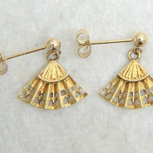 May include: A pair of gold-colored earrings. Each earring features a post with a small ball and a fan-shaped pendant with a cut-out design. The earrings are displayed on a light-colored surface.