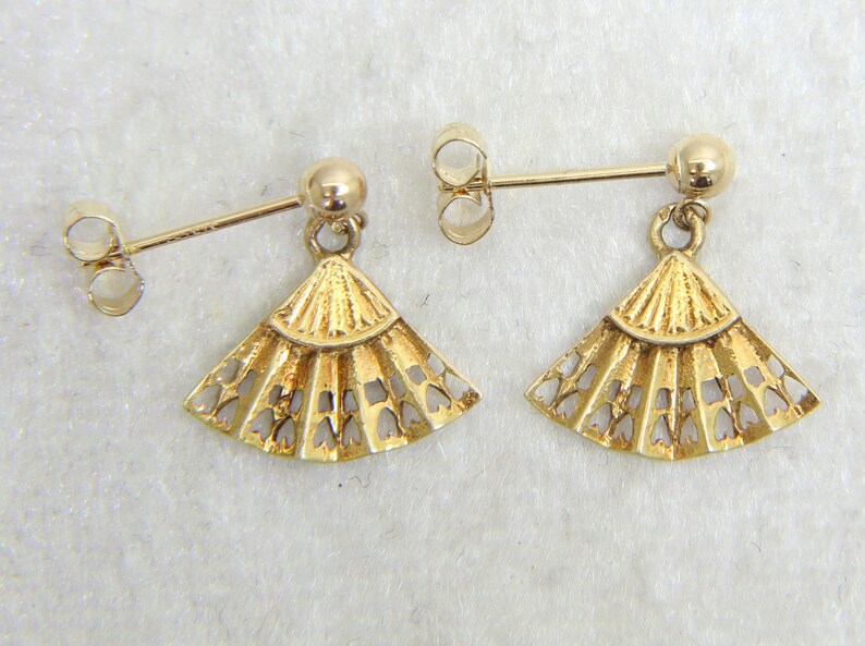 May include: A pair of gold-colored earrings. Each earring features a post with a small ball and a fan-shaped pendant with a cut-out design. The earrings are displayed on a light-colored surface.