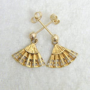 May include: A pair of gold-colored fan-shaped earrings. Each earring features a small, round stud and a delicate fan pendant with heart-shaped cutouts. The earrings are displayed against a white background.