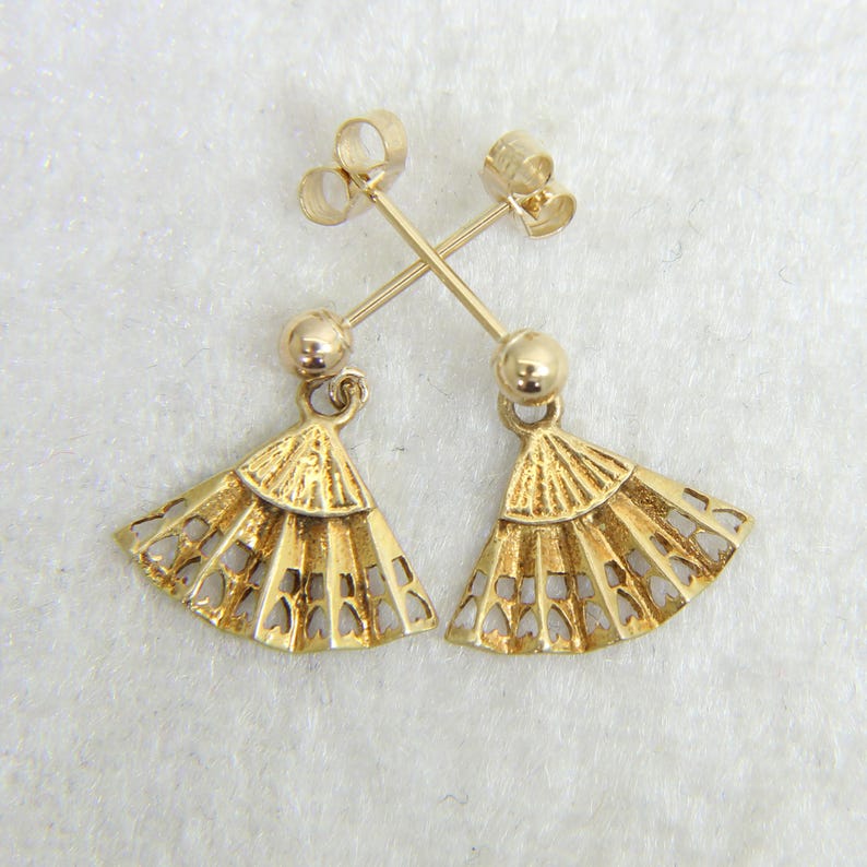 May include: A pair of gold-colored fan-shaped earrings. Each earring features a small, round stud and a delicate fan pendant with heart-shaped cutouts. The earrings are displayed against a white background.