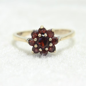 Può includere: Un anello dorato con un gruppo a forma di fiore di pietre preziose rosso scuro. La fascia dell'anello è sottile e le pietre preziose sono ravvicinate, creando un vivace motivo floreale. L'anello è posto su uno sfondo bianco morbido.