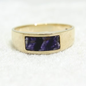 Puede incluir: Un anillo dorado con una piedra rectangular incrustada. La piedra presenta una mezcla de tonos morados oscuros y negros. El anillo tiene un diseño simple y clásico, adecuado para el uso diario.