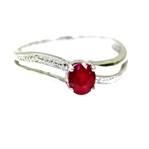 Puede incluir: Un anillo de plata con una piedra preciosa de rubí rojo engastada en el centro. El anillo tiene un diseño de doble banda con un patrón delicado.