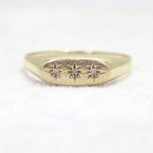 May include: A gold ring with a rectangular face. The face is adorned with three starburst patterns, each with a tiny, clear stone in the middle. The ring has a smooth, matte finish and is set against a white backdrop.