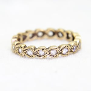 May include: A gold band ring with a delicate design featuring a row of small, clear gemstones set in a heart-shaped pattern.