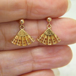 May include: A pair of gold-colored fan-shaped earrings. Each earring features a small, round stud at the top, connected to a delicate, openwork fan design. The earrings are held up by a person's fingers.