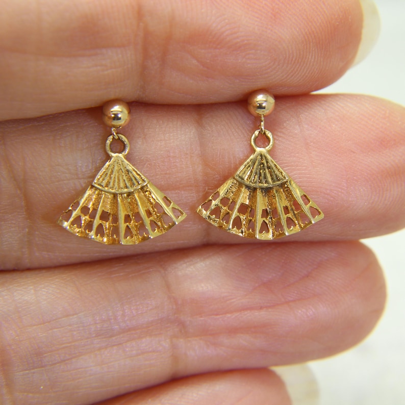 May include: A pair of gold-colored fan-shaped earrings. Each earring features a small, round stud at the top, connected to a delicate, openwork fan design. The earrings are held up by a person's fingers.
