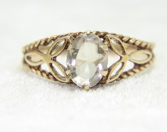 May include: A gold ring with an oval, clear gemstone. The ring has a decorative band with a braided design and openwork leaf-shaped accents. The gemstone is the focal point, set in a classic, elegant style.