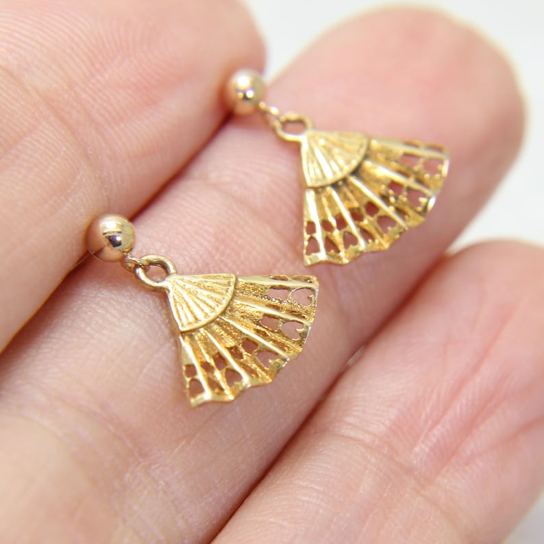 May include: A pair of gold-colored fan-shaped earrings. Each earring features a small, round stud and a delicate, openwork fan design. The earrings are made of a shiny, metallic material.