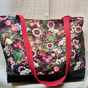 May include: A floral tote bag with a black base and bright red straps. The bag features a dark background covered in a colorful floral pattern with pink, white, and green flowers. The tote bag is rectangular and has two long straps.