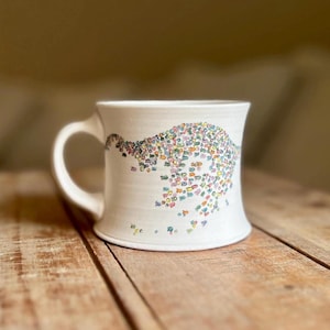 May include: White ceramic mug with a curved handle and a colorful, abstract design. The design features small, multi-colored shapes clustered together, resembling a wave or cloud. The mug sits on a wooden surface.