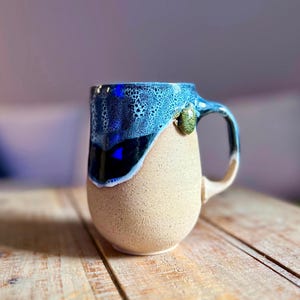 May include: A ceramic mug with a speckled beige base and a glossy, deep blue glaze dripping from the rim. A small, sculpted green turtle is attached to the side near the handle. The mug features a curved handle and a smooth, rounded body shape.
