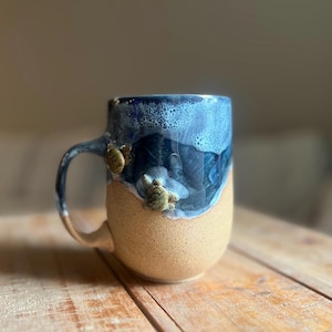 May include: Ceramic mug with a sea turtle theme. The mug features a tan base and a top section with a blue wave design. Two small sea turtle figurines are attached to the side of the mug. The handle is tan and matches the base.