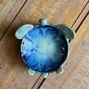 May include: A ceramic turtle-shaped dish featuring a deep blue and light blue glazed interior with a crackled texture. The outer rim and appendages are finished in a muted sage green glaze, highlighting the sculpted head, flippers, and tail.