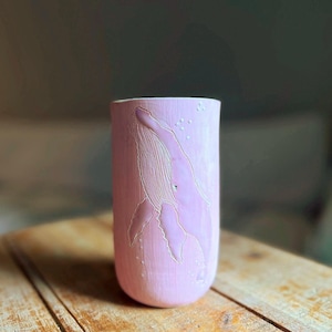 May include: A cylindrical ceramic tumbler in a soft pastel pink hue, featuring a detailed, etched relief of a humpback whale on its exterior surface. The whale design is highlighted with white outlines and small white dots representing bubbles.