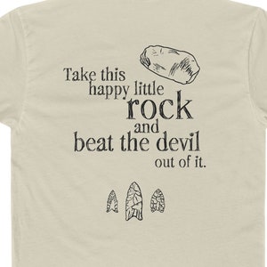May include: A beige t-shirt with the text "Take this happy little rock and beat the devil out of it." printed on the back. There are three small drawings of arrowheads below the text.