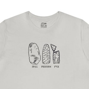 May include: A light gray t-shirt with a black and white illustration of three stone tools. The text "SPALL", "PRESHORM", and "F*CK" is printed below the tools.