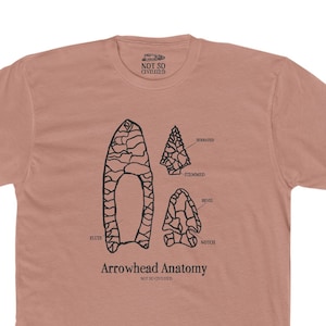 May include: A light brown t-shirt with a black illustration of an arrowhead and the text "Arrowhead Anatomy" printed on the front. The illustration shows the different parts of an arrowhead, including the flute, the stem, the shoulder, and the notch.