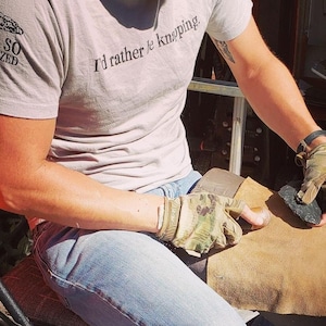 May include: A person wearing a grey t-shirt with the text "I'd rather be knapping" is sitting on a chair and working on a piece of flint with a leather pad.