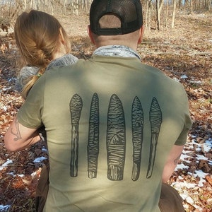 May include: A person wearing a green t-shirt with a graphic of five stylized arrows. The arrows are black and have a wood grain texture. The person is wearing a black baseball cap.