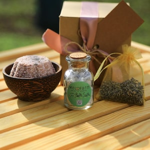 Op de afbeelding: Een cadeauset voor ontspanning in bad, met een bruine doos met een roze lint, een glazen pot met badzout met het label "Melofarm Bath Salts", een kleine kom met roze badzout en een klein zakje gedroogde lavendel.