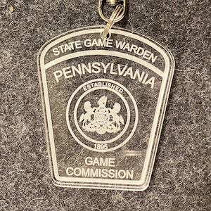 Puede incluir: Llavero de acrílico transparente con el emblema del Guardián del Juego del Estado de Pensilvania. El llavero tiene un cierre plateado y una forma de escudo con las palabras "State Game Warden", "Pennsylvania", "Established 1895" y "Game Commission".