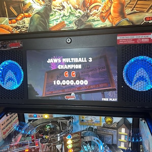 May include: A pinball machine with a digital display showing the game Jaws Multiball 3 Champion. The display shows a score of 10,000,000 and the text "Amity Island Welcomes You".