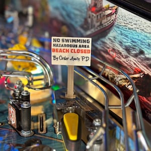 May include: A white sign with black text reads "NO SWIMMING HAZARDOUS AREA BEACH CLOSED By Order Amity PD". The sign is attached to a wooden stick and is placed on a pinball machine.