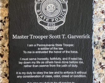 Könnte beinhalten: Schwarze Schiefertafel mit einem Abzeichen der Pennsylvania State Police und Text. Die Tafel ist mit dem Namen "Master Trooper Scott T. Garverick" und einer Widmung graviert. Die Tafel misst ca. 20 x 25 cm.