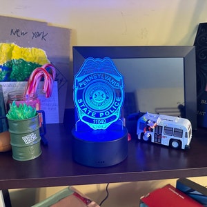 May include: A blue LED night light shaped like a Pennsylvania State Police badge. The badge is illuminated with blue light and features the text "PENNSYLVANIA STATE POLICE" and "11040". The light sits on a black base. Other items on the shelf include a toy bus and a framed photo.