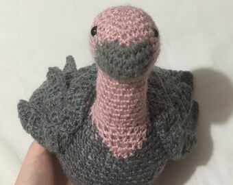 May include: A crocheted stuffed bird with a pink head and neck and gray body. The bird has black eyes and a gray beak.