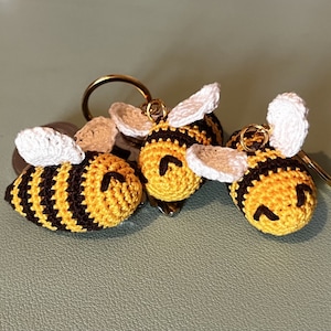 May include: Three crocheted bee keyrings with black and yellow stripes, white wings, and black eyes. The keyrings are attached to gold key rings.