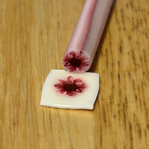 May include: A close-up of a polymer clay cane and a square tile, both featuring a dark red flower design. The cane is pink and white, with the flower visible at the end. The tile is white with the flower in the center.