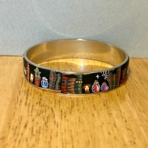 May include: A silver-toned metal bangle bracelet featuring a black enamel inlay decorated with a repeating pattern of colourful books, potted plants, glowing candles, and small glass potion bottles in various shades of red, blue, and purple.