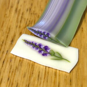 May include: A polymer clay cane featuring a detailed purple and white floral design with green leaves, shown as a long cylindrical log and a single thin, flat slice displaying the intricate lavender-like flower pattern on a white background.