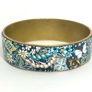 Pode incluir: Um bracelete dourado com um design floral azul, branco e verde. O design apresenta flores, borboletas e folhas.