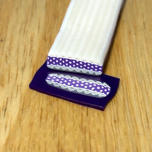 May include: A white rectangular polymer clay piece with a purple and white polka dot design, accented with white lace trim, rests on a purple base. The design is repeated on the base. The background is a wooden surface.