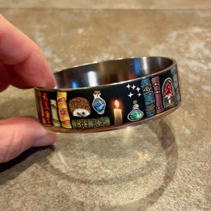 May include: A silver-toned metal bangle bracelet featuring a black wrap-around design. The graphic print displays a row of colorful books, a small hedgehog, glowing potion bottles, a lit candle, and a mushroom inside a glass dome.