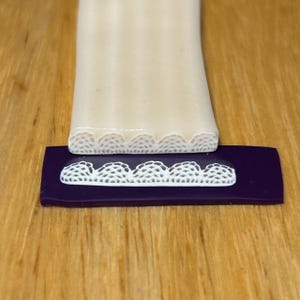 Puede incluir: Primer plano de un sello con patrón de encaje blanco sobre una base púrpura, con un patrón de encaje blanco a juego en la parte inferior de un objeto rectangular color crema. Los objetos están sobre una superficie de madera.