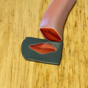 May include: A close-up of polymer clay art. A light pink clay tube with a red leaf design on the end is positioned above a gray clay piece with a matching leaf design. The background is a light brown wooden surface.