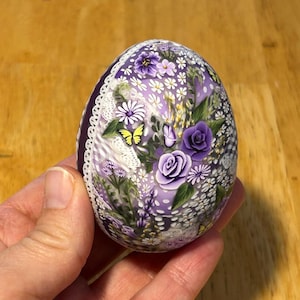 May include: A decorative egg featuring a floral pattern. The egg is adorned with purple roses, white daisies, and yellow butterflies. A white lace trim edges the egg. The egg is held in a person's hand.