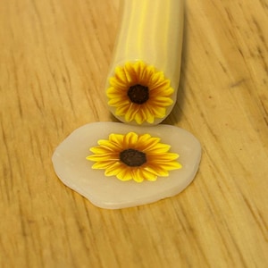 May include: A cylindrical polymer clay cane featuring a sunflower pattern with yellow petals and a dark brown center, positioned above a single, thin, circular slice of the same sunflower design set against a translucent, off-white clay base.