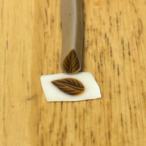 May include: A brown leaf stamp is pressed onto a white surface, creating a detailed leaf impression. The stamp is a light brown color and has a leaf design on the end. The background is a light brown wooden surface.