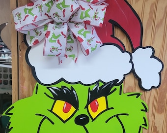 Puede incluir: Una cara verde del Grinch con ojos rojos y un gorro de Papá Noel blanco con un borde rojo y un lazo blanco con un patrón verde y blanco. El lazo tiene un fondo blanco con piernas verdes del Grinch y bastones de caramelo rojos.