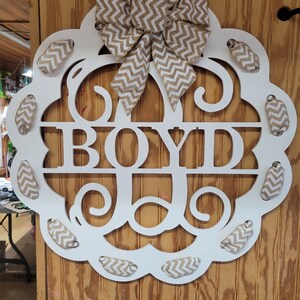 May include: White wooden monogram door hanger with the last name "BOYD" in large letters. The monogram is decorated with a brown and white chevron patterned burlap bow and smaller burlap bows around the edge.