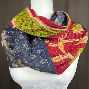 May include: A quilted infinity scarf featuring a patchwork design with sections of lime green fabric with brown floral patterns, deep red fabric with yellow leaf motifs, and dark navy blue fabric with a subtle ikat-style pattern and white stitching.