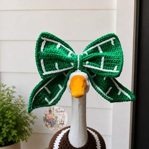 May include: A large, green, crocheted bow with white stitched details worn on the head of a white goose figurine. The goose has an orange beak and is wearing a brown crocheted garment with a white trim around its neck and chest area.