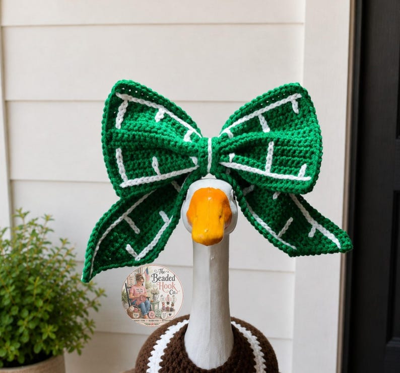May include: A large, green, crocheted bow with white stitched details worn on the head of a white goose figurine. The goose has an orange beak and is wearing a brown crocheted garment with a white trim around its neck and chest area.